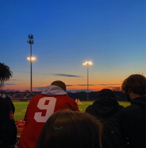 Students gather in the stands to watch a boys lacrosse match on the turf at sunset. 
